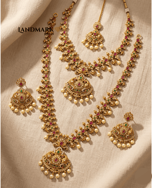 Landmark Jewels bridal jewelry set featuring long and short antique gold necklaces, earrings, and Maang Tikka with red/green stones and pearls.
