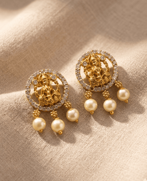 Pair of Landmark Jewels gold-plated temple stud earrings featuring a central Goddess Lakshmi motif surrounded by a circular border of white CZ stones and finished with three hanging pearls on a textured beige fabric background.