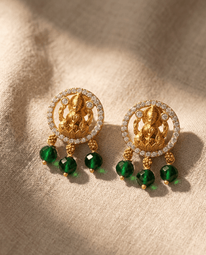 Gold-plated circular frame earrings lined with white stones surrounding a central Goddess Lakshmi motif, featuring three hanging emerald-green faceted beads.