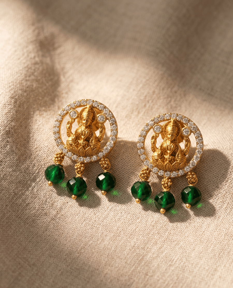 gold-plated-circular-frame-earrings-lined-with-white-stones-surrounding-a-central-goddess-lakshmi-motif-featuring-three-hanging-emerald-green-faceted-beads