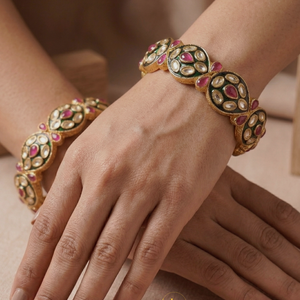 Pair of gold-plated Indian bangles with ruby stones, white polki crystals, and black enamel work on a floral background.