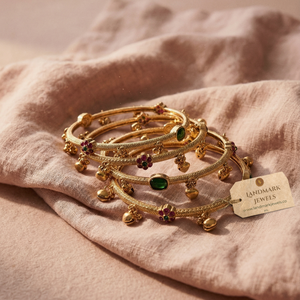 Antique gold-plated bangles with ruby and emerald floral kemp stones and small hanging golden bells on a peach fabric background.