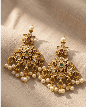 Pair of Landmark Jewels antique gold-plated Jhumka earrings featuring intricate floral metalwork, white Kundan stones, green and red accents, and hanging pearl drops against a textured beige fabric backdrop.