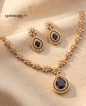 Landmark Jewels gold-toned necklace and earring set featuring pear-shaped blue sapphire-style stones and white zircon floral accents on a beige fabric background.