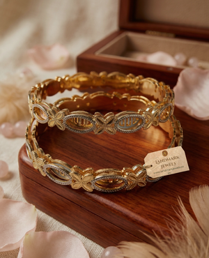 Set of two gold and silver dual-tone bangles with butterfly and oval filigree patterns resting on a wooden jewelry box.