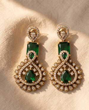 A pair of elegant Landmark Jewels matte gold drop earrings featuring rectangular and tear-drop shaped emerald green stones surrounded by sparkling cubic zirconia accents, resting on a beige fabric background.