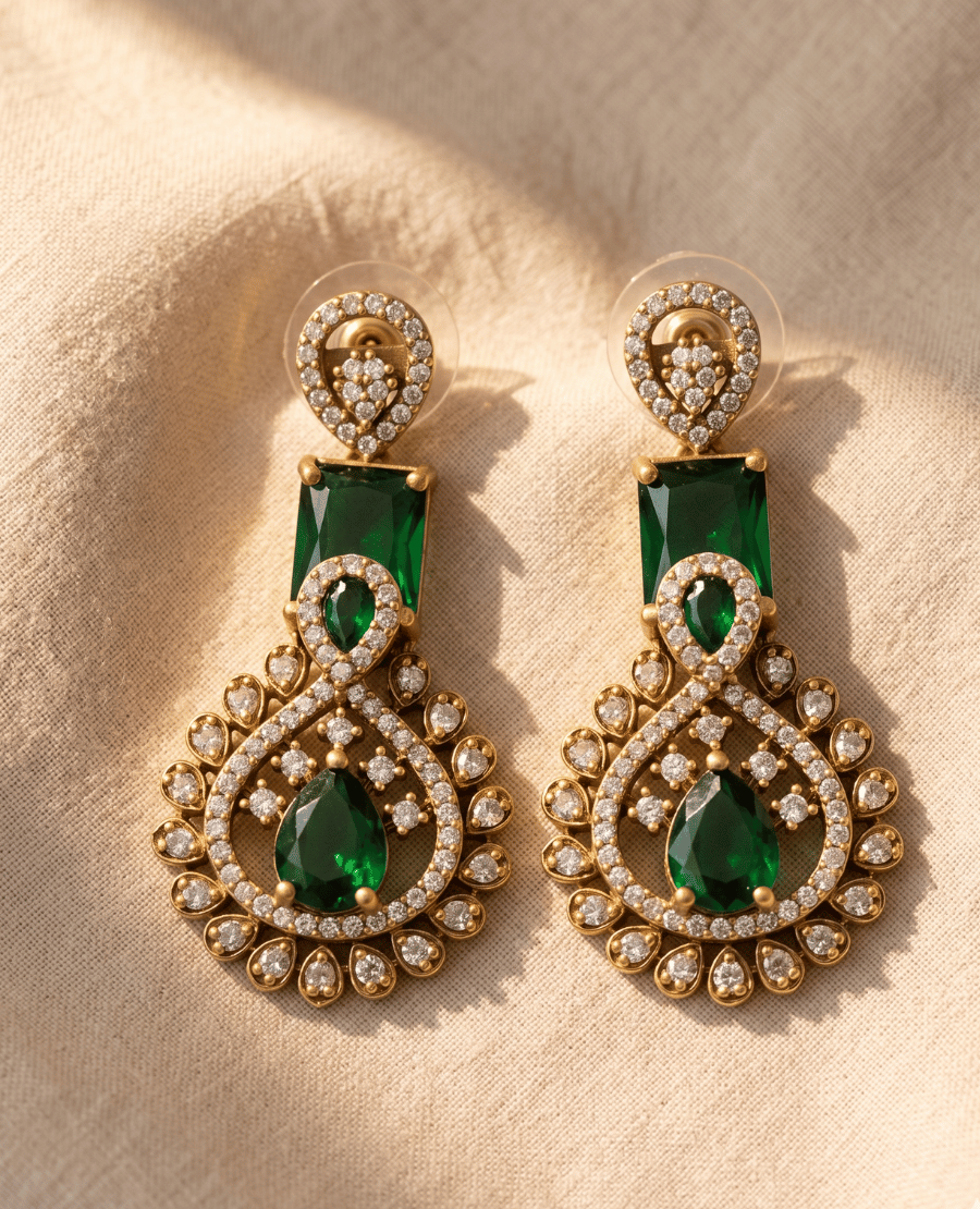 a-pair-of-elegant-landmark-jewels-matte-gold-drop-earrings-featuring-rectangular-and-tear-drop-shaped-emerald-green-stones-surrounded-by-sparkling-cubic-zirconia-accents-resting-on-a-beige-fabric-background