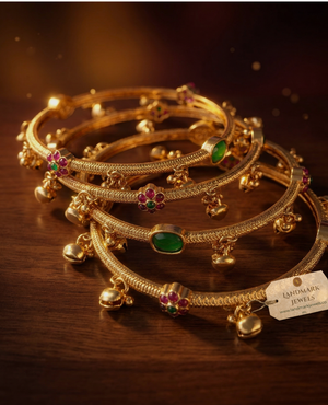 Antique gold-plated bangles with ruby and emerald floral kemp stones and small hanging golden bells on a peach fabric background.