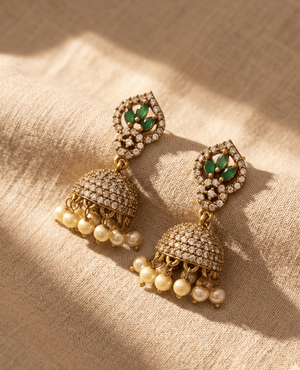 Elegant gold Jhumka earrings with a leaf-shaped top featuring three green marquise stones, a crystal-paved dome, and dangling cream-colored pearls.