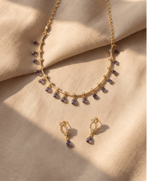 Gold chain necklace with spaced white crystals and dangling blue-purple tear-drop stones, with matching earrings on a neutral backdrop.