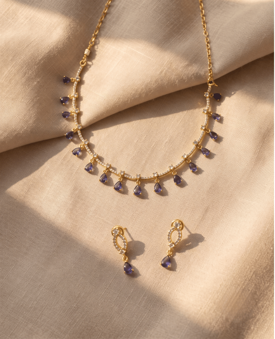 gold-chain-necklace-with-spaced-white-crystals-and-dangling-blue-purple-tear-drop-stones-with-matching-earrings-on-a-neutral-backdrop