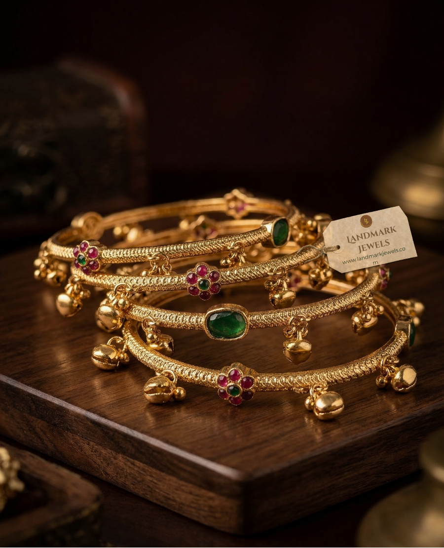 antique-gold-plated-bangles-with-ruby-and-emerald-floral-kemp-stones-and-small-hanging-golden-bells-on-a-peach-fabric-background