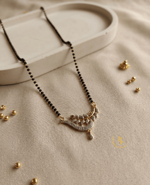 Landmark Jewels imitation mangalsutra with a single strand of black beads and a gold-plated pendant featuring a sparkling CZ-encrusted leaf and vine design on a cream fabric background.