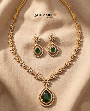 Landmark Jewels gold-plated necklace set featuring a large emerald green teardrop pendant and matching floral drop earrings on a beige fabric background.