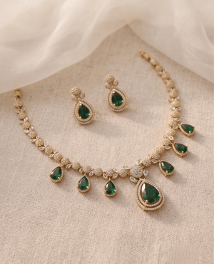 Gold necklace and earrings set featuring dangling green pear-shaped emerald stones and sparkling crystals on a neutral fabric background.