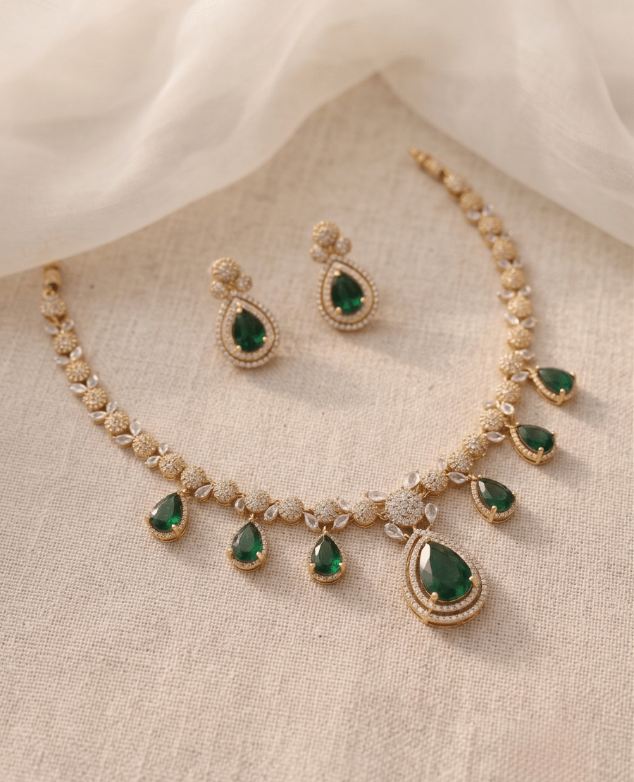 gold-necklace-and-earrings-set-featuring-dangling-green-pear-shaped-emerald-stones-and-sparkling-crystals-on-a-neutral-fabric-background