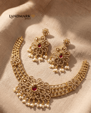 Landmark Jewels gold-plated choker necklace and earring set featuring floral patterns, ruby-red teardrop stones, cubic zirconia embellishments, and hanging pearl drops on a beige fabric background.