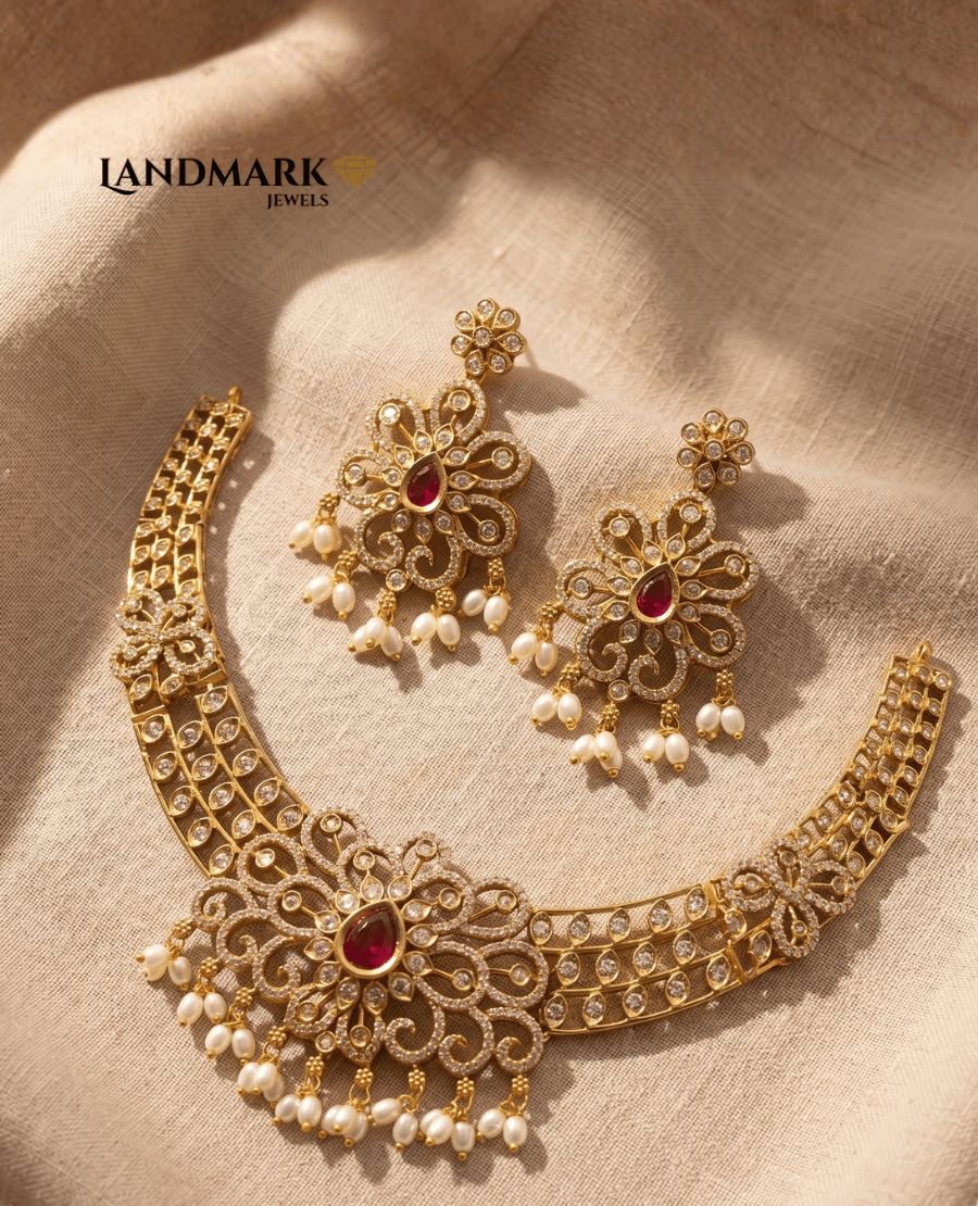 landmark-jewels-gold-plated-choker-necklace-and-earring-set-featuring-floral-patterns-ruby-red-teardrop-stones-cubic-zirconia-embellishments-and-hanging-pearl-drops-on-a-beige-fabric-background
