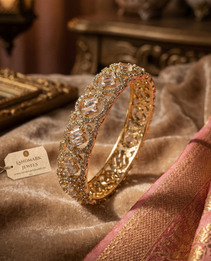 A pair of gold-plated American Diamond bangles featuring circular patterns and central baguette-cut stones, resting on a soft pink velvet surface with a Landmark Jewels brand tag.