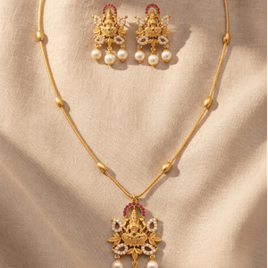 Matte gold temple necklace with a Goddess Lakshmi pendant framed by red stones and pearls, alongside matching Lakshmi stud earrings.