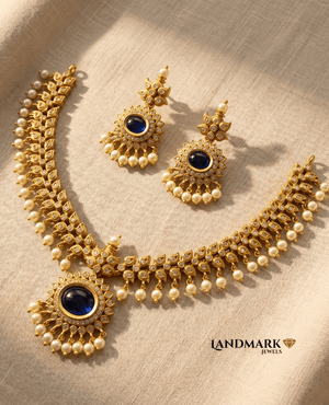 Landmark Jewels antique gold necklace and earring set featuring central royal blue kemp stones, white crystals, and dangling pearls on a beige fabric background.
