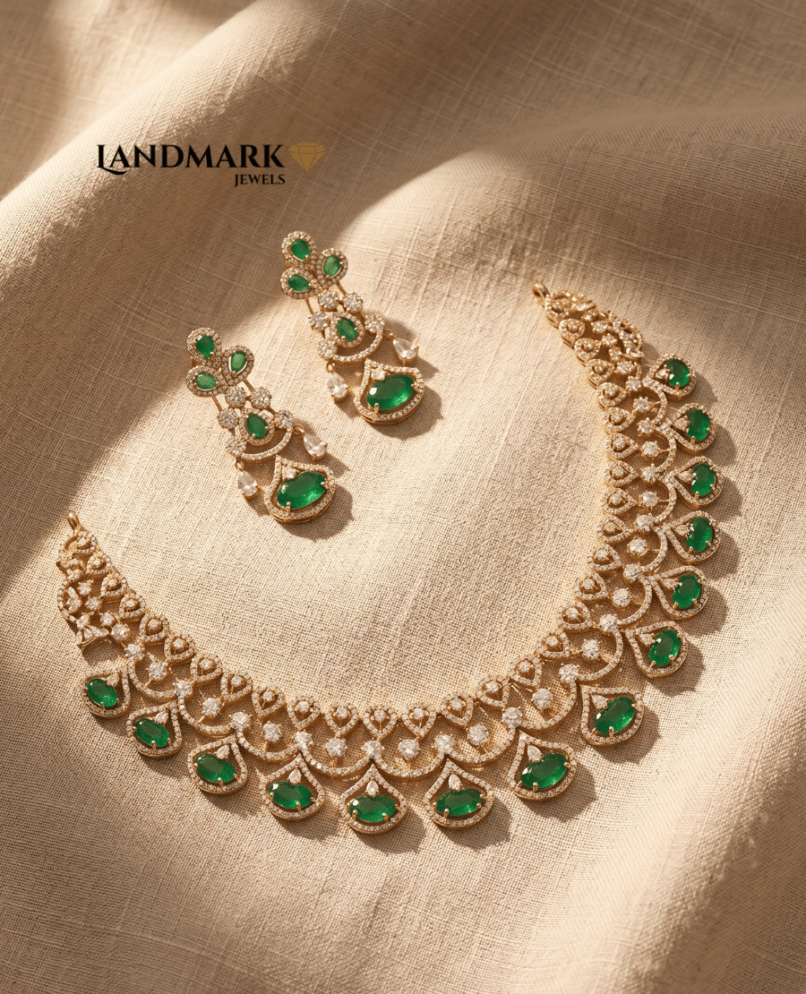 landmark-jewels-imitation-jewelry-set-featuring-a-gold-plated-choker-and-matching-drop-earrings-studded-with-emerald-green-stones-and-american-diamonds-on-a-beige-fabric-background