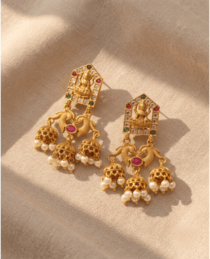Traditional antique gold temple jhumka earrings featuring Goddess Lakshmi, ruby stones, and hanging pearls on a neutral fabric background.