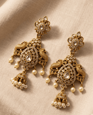 Heavy statement antique gold earrings featuring dual peacock motifs, a large center teardrop stone, hanging pearl beads, and a small central dangling jhumka.