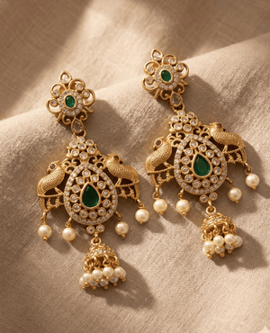Intricate antique gold chandelier earrings featuring dual peacock motifs flanking a teardrop emerald-green stone, adorned with hanging pearls and a small central jhumka bell.