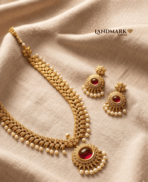 Landmark Jewels antique gold-plated necklace and earring set featuring deep red ruby stones, intricate leaf patterns, and white pearl droplet accents resting on a beige linen fabric.