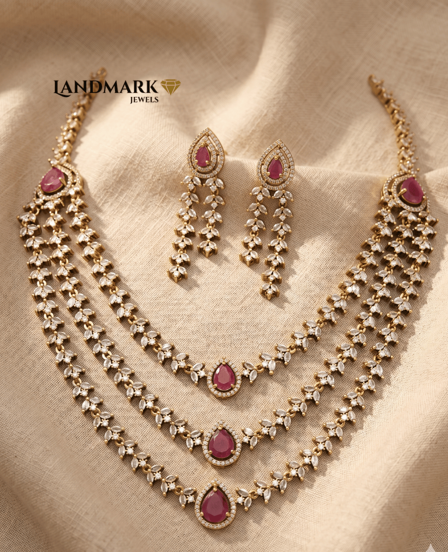 landmark-jewels-3-layered-gold-toned-necklace-and-earring-set-featuring-white-american-diamonds-and-pear-shaped-red-ruby-accents-on-a-beige-fabric-background