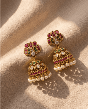 Vibrant temple-style gold Jhumka earrings featuring a lotus-shaped top with red and green kemp stones, a decorated dome, and small hanging white pearls casting a shadow on fabric.