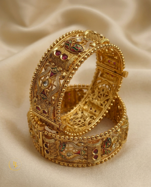 Pair of antique gold-plated Meenakari Kada bangles with green and red enamel work and ruby stone accents on a beige silk background.