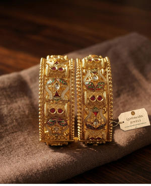 Pair of antique gold-plated Meenakari Kada bangles with green and red enamel work and ruby stone accents on a beige silk background.