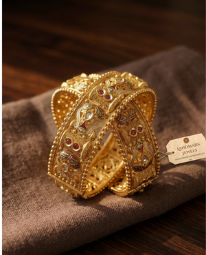 Pair of antique gold-plated Meenakari Kada bangles with green and red enamel work and ruby stone accents on a beige silk background.