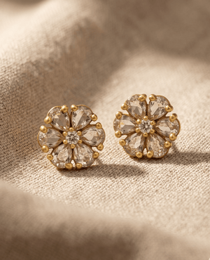 Pair of matte gold-plated floral stud earrings with clear teardrop crystals on a textured beige fabric background.