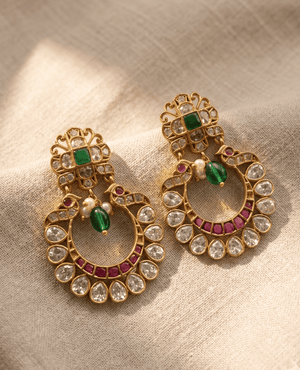 Round gold-plated chandbali earrings featuring a peacock design, white stones, ruby lining, and a central green teardrop bead.