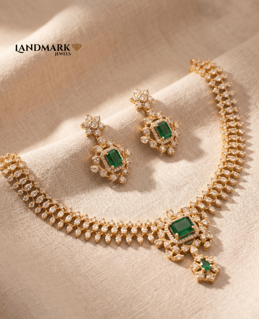 landmark-jewels-imitation-jewelry-set-featuring-a-gold-toned-necklace-with-emerald-cut-green-stones-and-leaf-shaped-diamond-accents-accompanied-by-matching-floral-halo-earrings-on-a-neutral-fabric-background