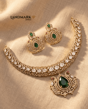 Landmark Jewels gold-plated choker necklace and earring set featuring emerald green pear-cut stones, white polki-style gems, and intricate peacock engravings on a beige fabric background.
