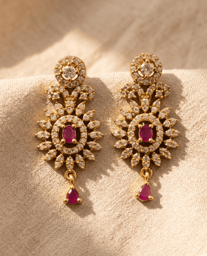 Ornate gold-tone long drop earrings featuring a sunburst design with white crystals, oval ruby centers, and a single hanging ruby teardrop at the bottom.