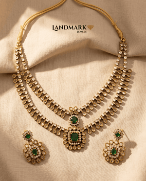 Landmark Jewels imitation jewelry set featuring a gold-toned dual-layered necklace with white teardrop stones, emerald green center pendants, and matching floral drop earrings on a neutral fabric background.