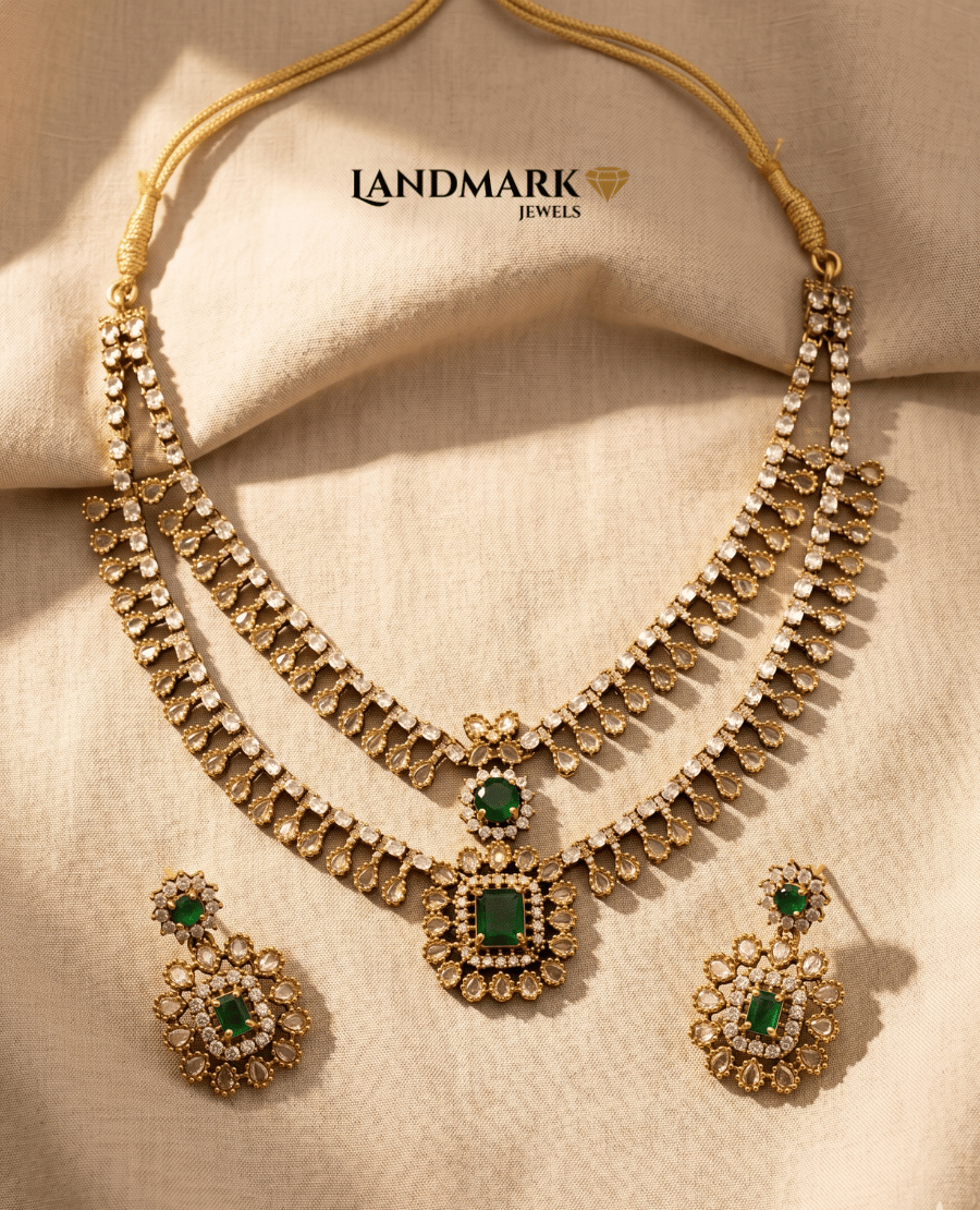 landmark-jewels-imitation-jewelry-set-featuring-a-gold-toned-dual-layered-necklace-with-white-teardrop-stones-emerald-green-center-pendants-and-matching-floral-drop-earrings-on-a-neutral-fabric-background