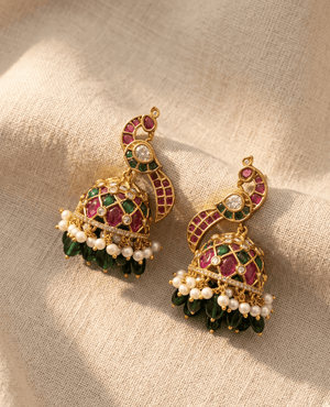 Royal peacock design gold-plated jhumka earrings inlaid with pink and green stones, finished with white pearls and dark green drop beads.