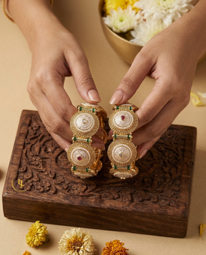 Pair of antique gold-plated Indian kada bangles with white Meenakari enamel work, pearls, and red stones resting on a carved wooden box with floral background