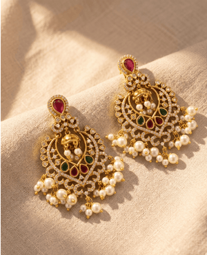Grand gold-plated crescent chandbali earrings featuring brilliant cubic zirconia, red and green accent stones, and heavy pearl cluster dangles.