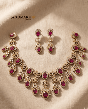 Landmark Jewels imitation jewelry set featuring a gold-plated necklace and drop earrings adorned with oval ruby-red stones and white sparkling crystals on a cream fabric background.