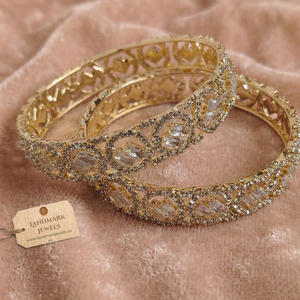 A pair of gold-plated American Diamond bangles featuring circular patterns and central baguette-cut stones, resting on a soft pink velvet surface with a Landmark Jewels brand tag.