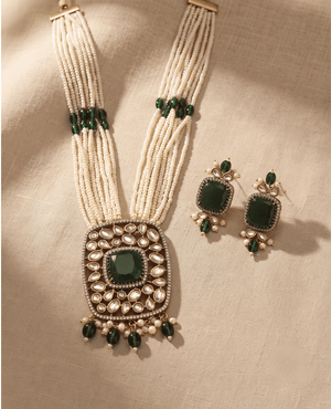 Multi-strand white seed bead necklace with a large rectangular pendant featuring a central green stone and Polki work, alongside matching earrings.