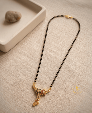 Landmark Jewels gold-plated short mangalsutra with floral American Diamond pendant, green and pink stones, and traditional black beads on a beige fabric background.