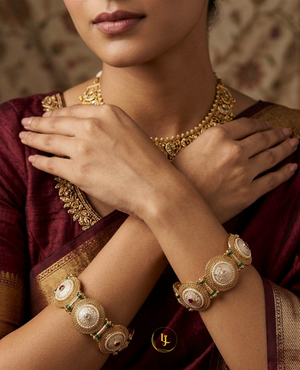Pair of antique gold-plated Indian kada bangles with white Meenakari enamel work, pearls, and red stones resting on a carved wooden box with floral background